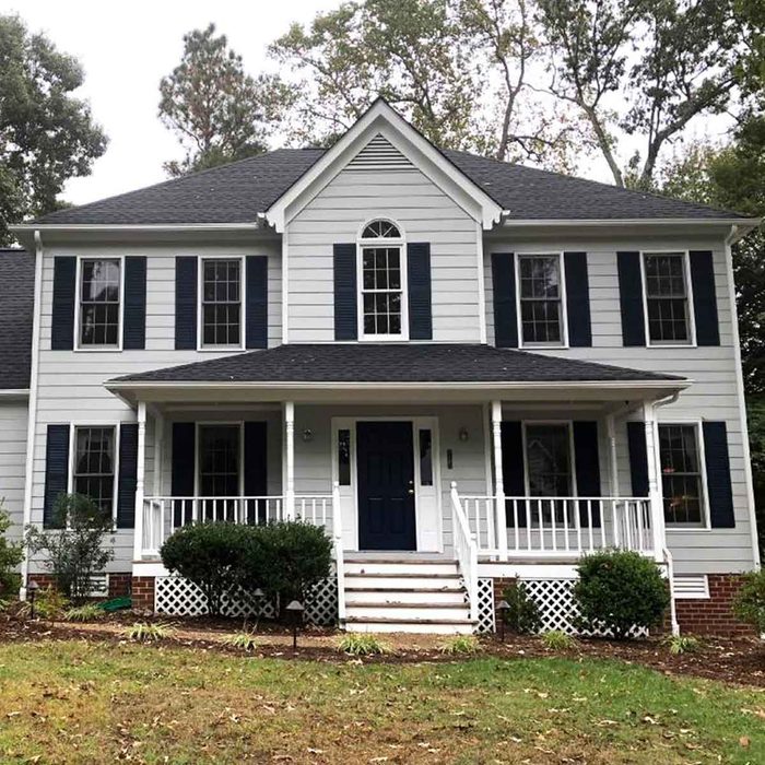 A two-story, light gray house with dark green shutters and a dark door sits on a grassy yard, surrounded by trees and shrubs.