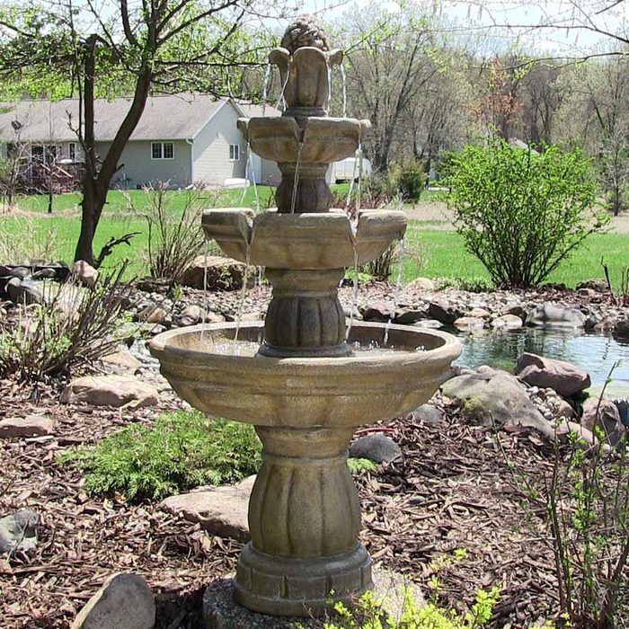 A stone fountain releases water from multiple tiers, surrounded by greenery and rocks, near a pond and a house in a sunny outdoor setting.