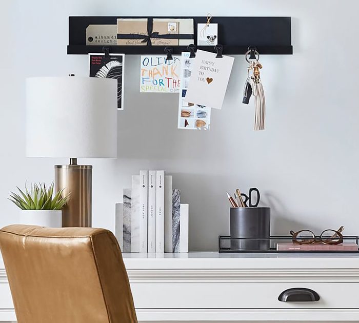 A desk holds a lamp and books. A shelf above displays notes and clips. A chair is positioned nearby, creating a cozy workspace atmosphere.