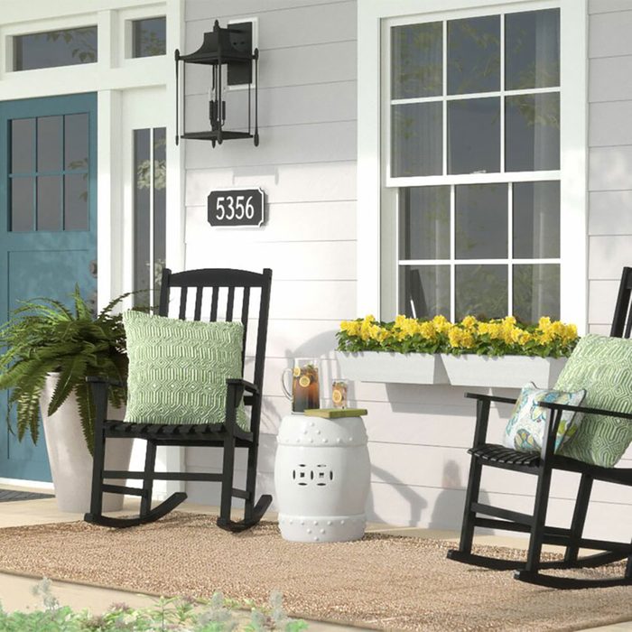 Two rocking chairs with decorative pillows sit on a porch, accompanied by a white garden stool and a planter, with a house number and flower box nearby.