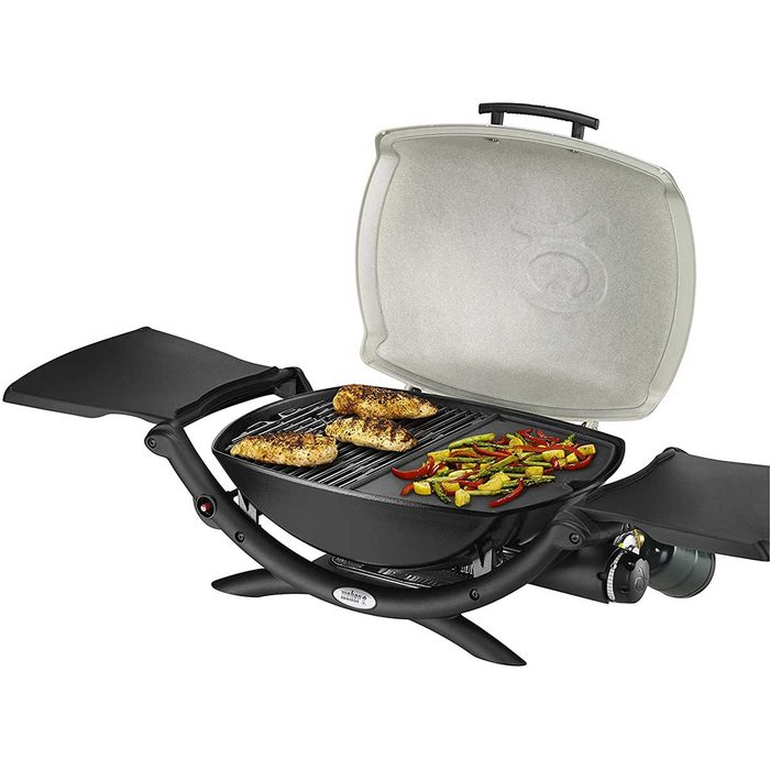 A portable grill with an open lid cooks seasoned chicken and colorful vegetables on its grates, set against a neutral background.