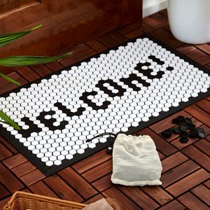 A black-and-white hexagonal welcome mat sits on wooden flooring, with a small mesh bag and scattered black pieces nearby, surrounded by a plant.