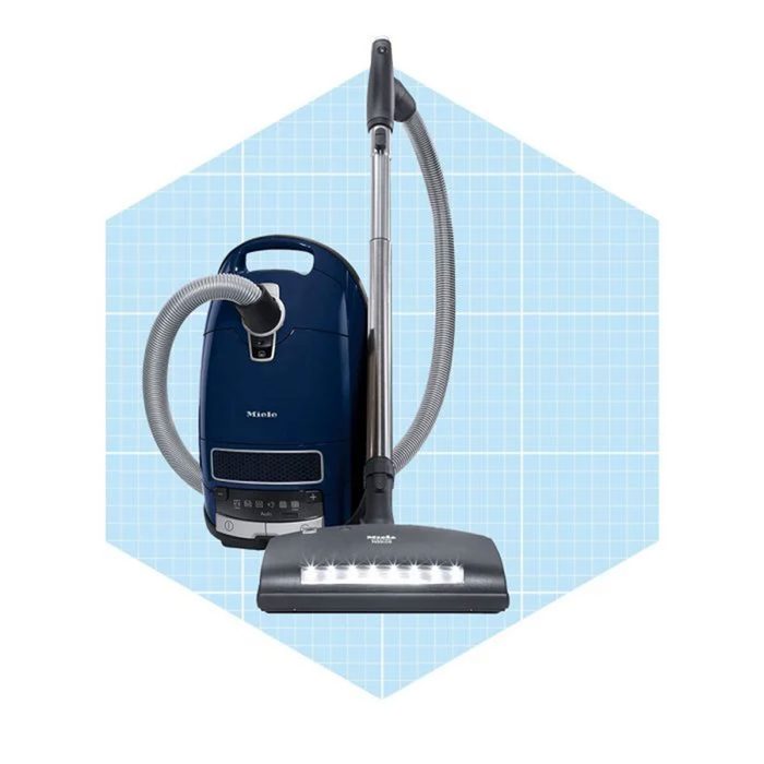 A blue canister vacuum cleaner stands upright with a silver hose and attachment, set against a light blue grid-patterned background.