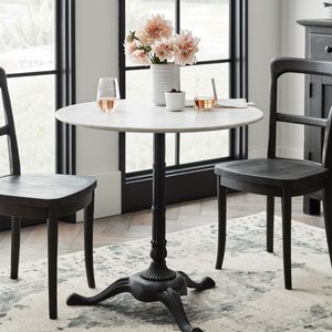 A round marble table with flowers sits between two black chairs in a bright room featuring large windows and a decorative rug.