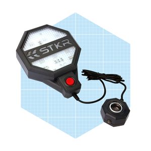 A portable LED work light illuminates a surface, connected by a wire to a round base, positioned on a grid background.