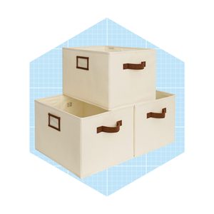 Three fabric storage bins, stacked with the top bin resting on two below, feature brown handles. They stand against a light blue grid background.