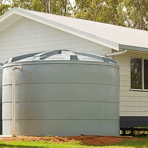 A large, cylindrical water tank sits beside a house, collecting rainwater under a clear sky, with greenery visible in the background.