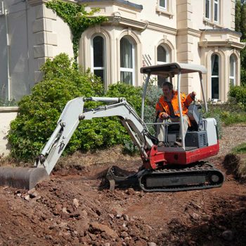 A small excavator digs earth in a garden while an operator, wearing an orange jacket, maneuvers it next to a large house and tools.
