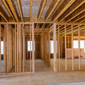 Wooden framing structures stand upright inside an under-construction building, with large windows allowing light in and a wooden floor beneath.