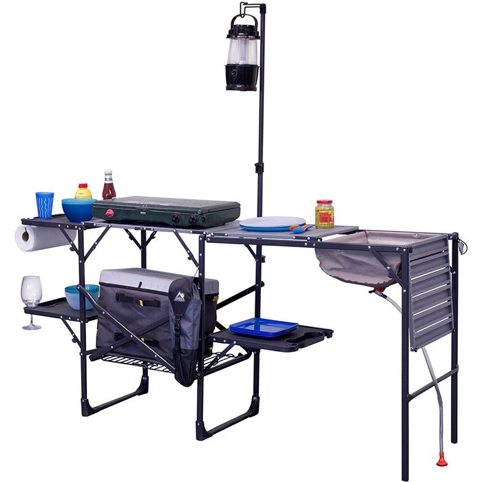 A camping kitchen setup displays a portable table with cooking equipment, condiments, dishes, and a lantern, organized for outdoor food preparation.