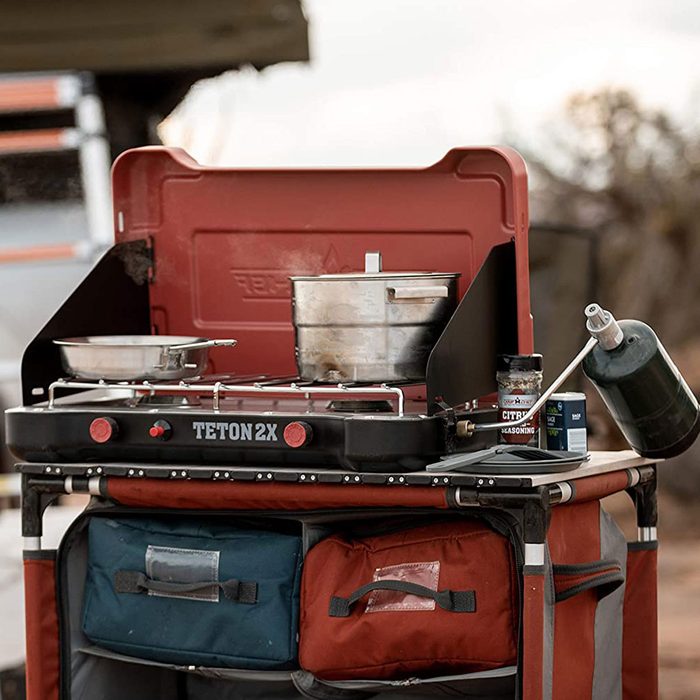 A portable camping stove with pots is set up for cooking outdoors, alongside containers and a gas canister, against a natural backdrop.