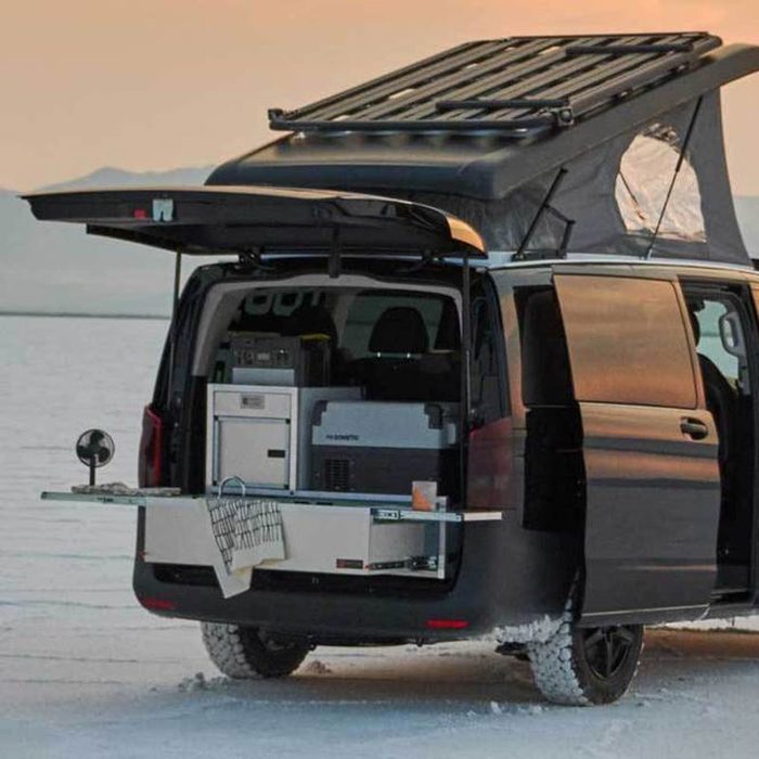 A black van with an elevated roof is parked on a snowy surface, displaying an open trunk with a kitchen setup and storage compartments visible.