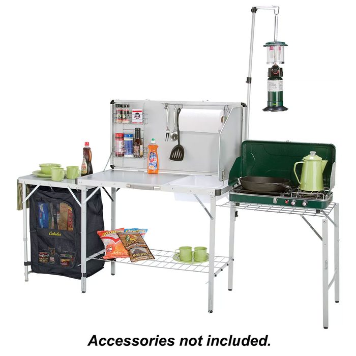 A folding camp kitchen features a countertop with cooking supplies and an attached gas stove, set in an outdoor setting. Accessories not included.