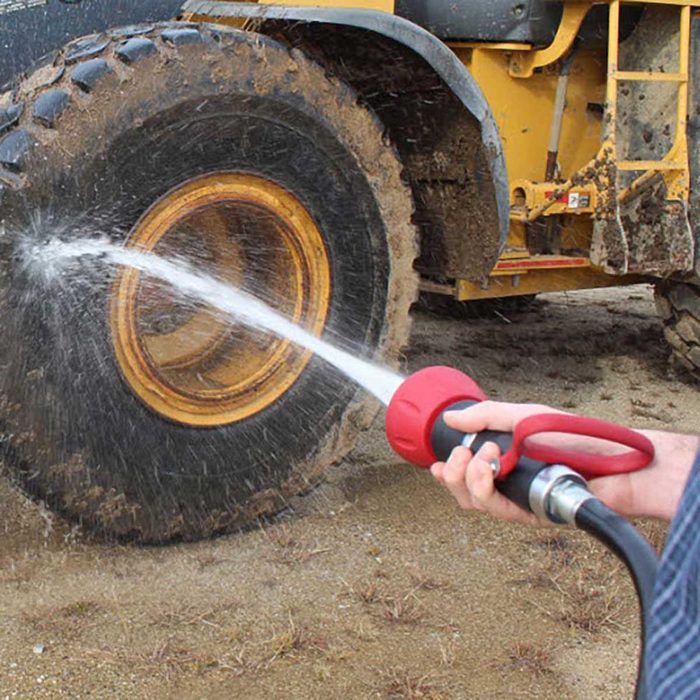 A person is using a hose to spray water on a dirty machine tire, cleaning it in a sandy environment.