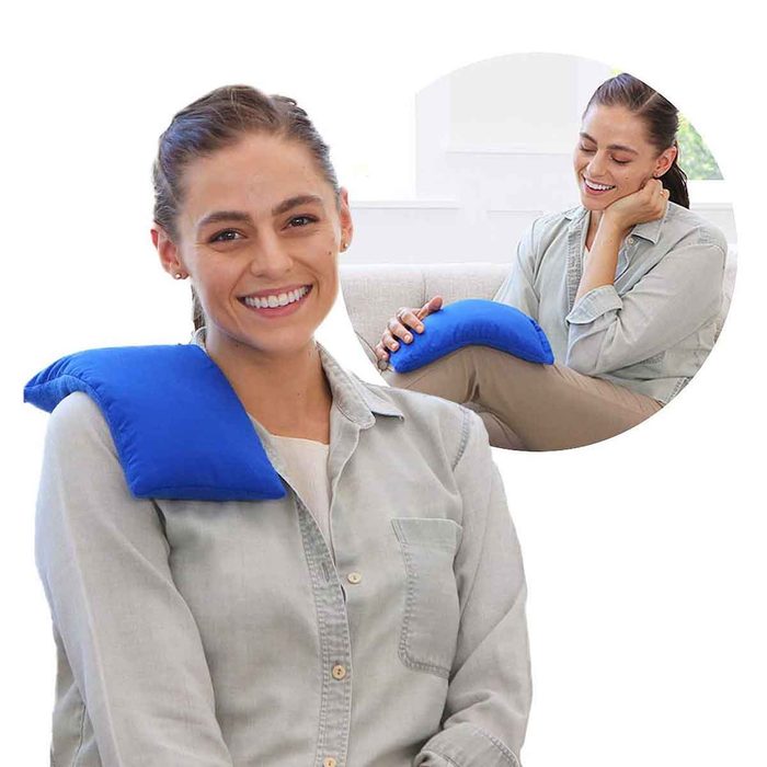 A smiling woman sits comfortably on a couch, wearing a shirt with a blue heating pad on her shoulder, indicating relaxation and relief.