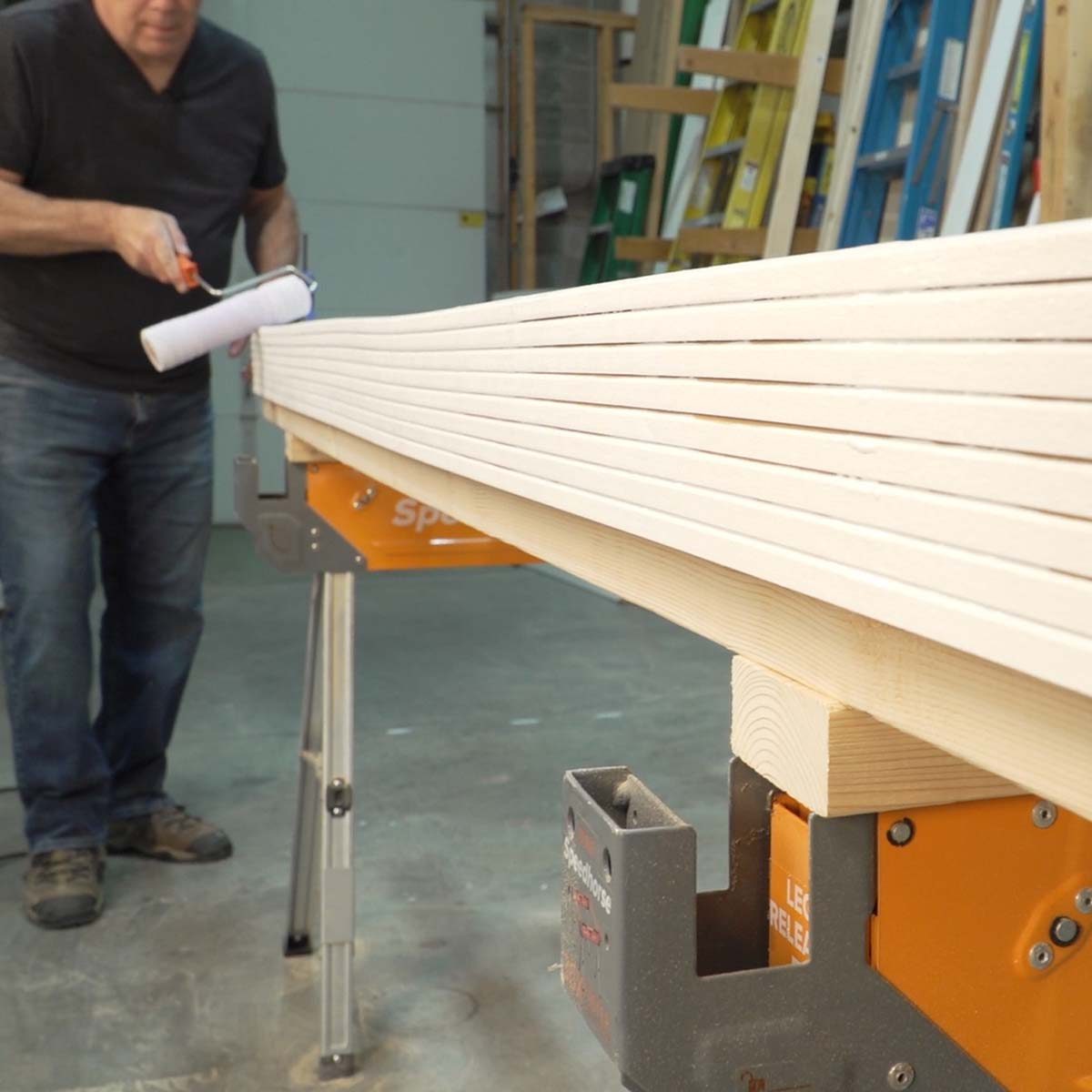 A person rolls paint on stacked wooden planks, using a paint roller in a workshop with ladders and equipment in the background.