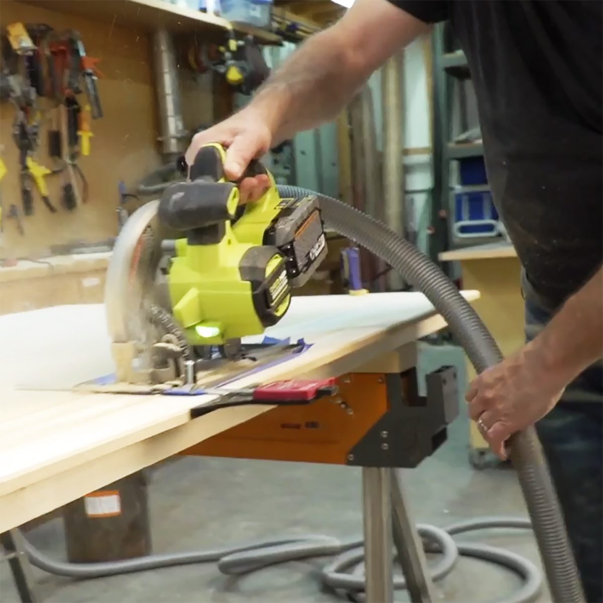 A person uses a yellow cordless circular saw to cut wood on a workbench, while dust collects in the air in a workshop environment.