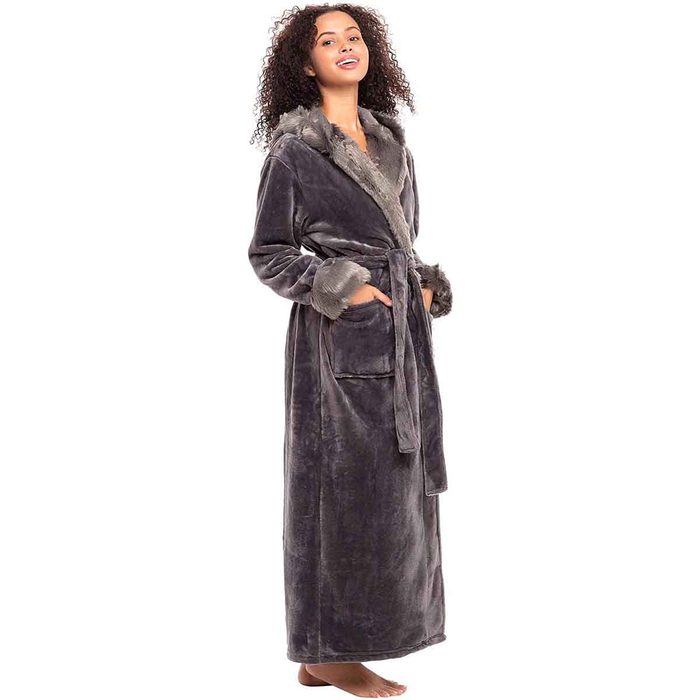 A woman stands and smiles, wearing a long, gray robe with a fur collar and cuffs, evoking a cozy, comfortable atmosphere.