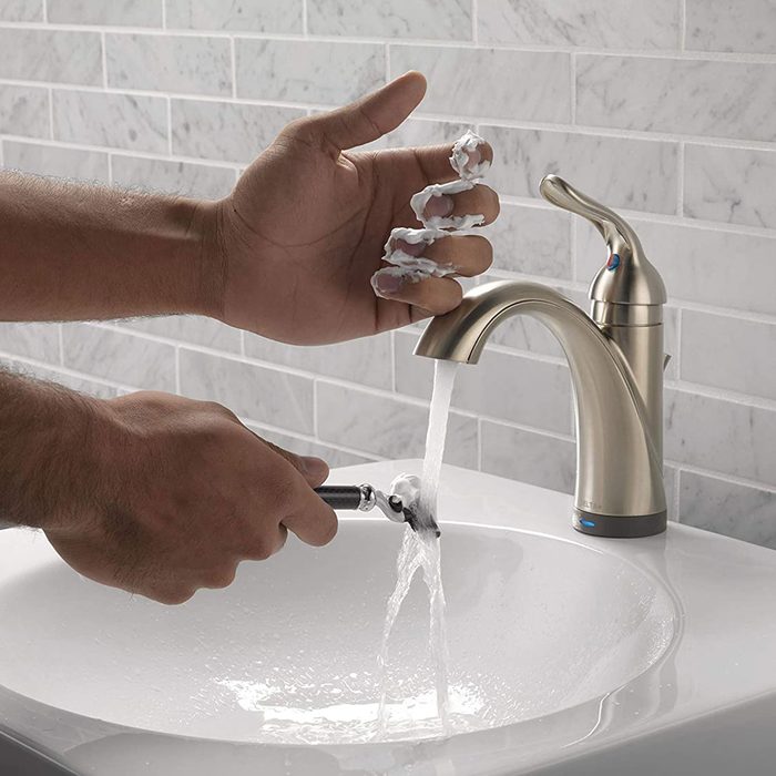 A hand applies shaving cream while holding a razor under running water from a faucet, positioned above a sink with a marble tile backdrop.