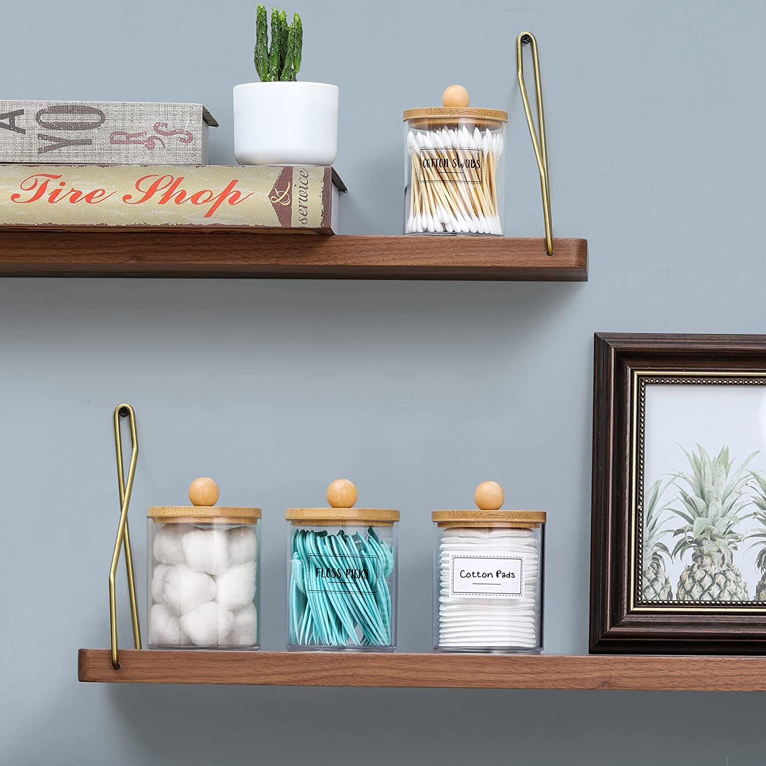 Clear jars filled with cotton balls, swabs, and pads are arranged on wooden shelves alongside books and a plant against a gray wall.