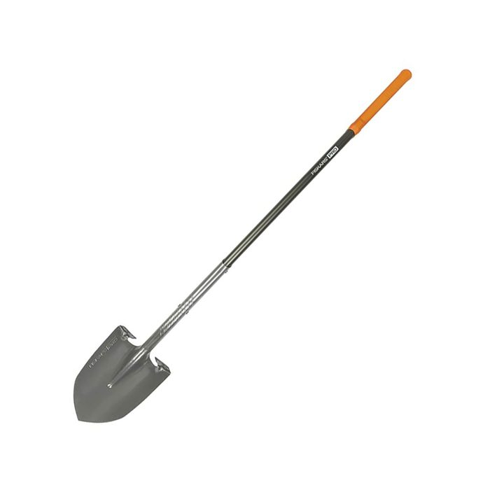 A metal shovel stands upright, featuring a pointed blade and a long handle with an orange grip, typically used for digging in soil or sand.