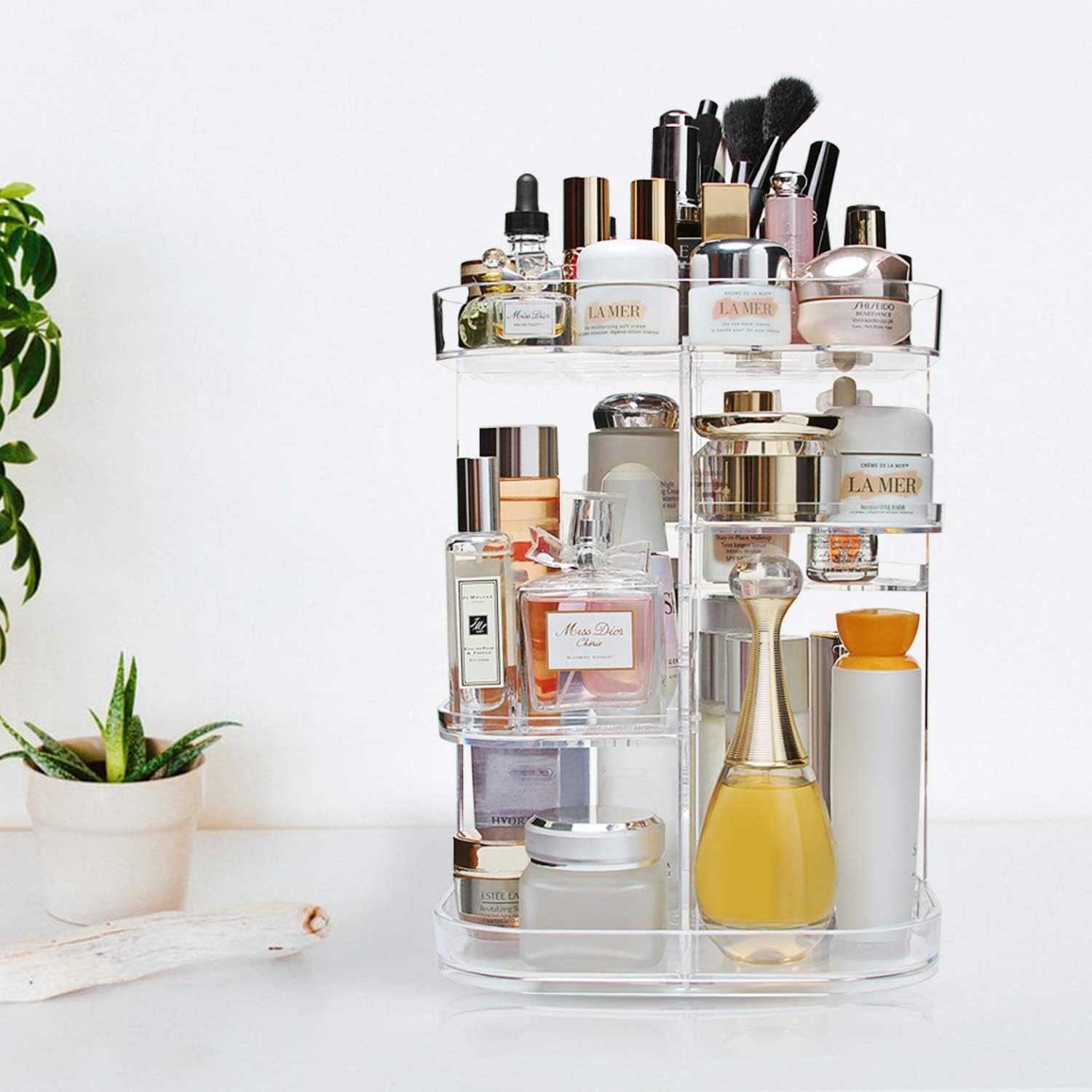 A clear organizer holds various skincare and fragrance products, positioned on a white surface next to a small plant, enhancing an elegant, tidy environment.