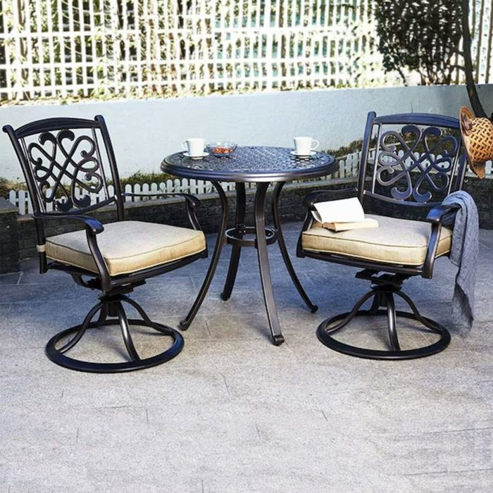 Two swivel chairs and a round table sit on a paved surface, surrounded by greenery and a white fence, with two cups and a small dish on the table.