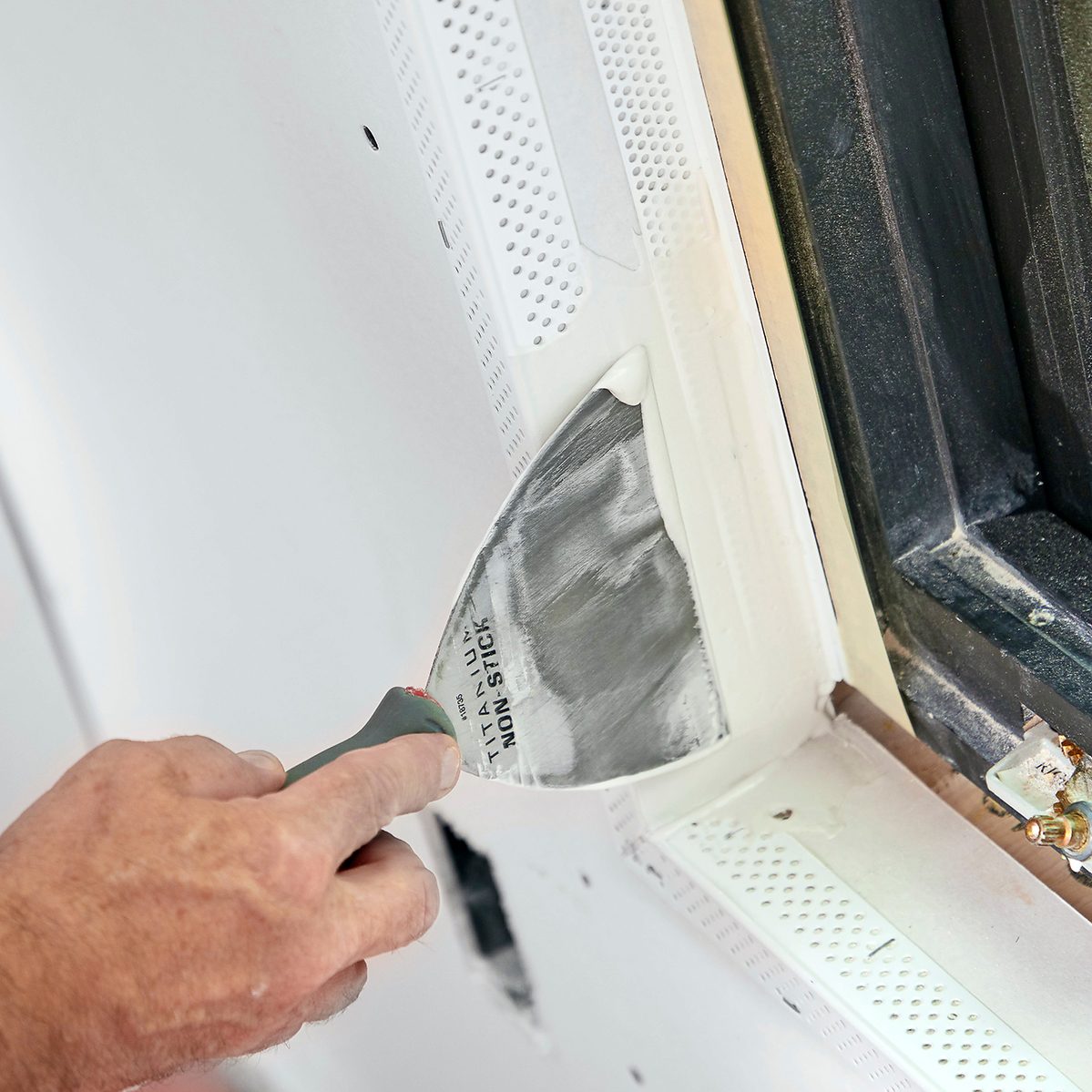 A hand holds a putty knife, applying compound to a drywall corner near a window frame, in a light-colored interior space.