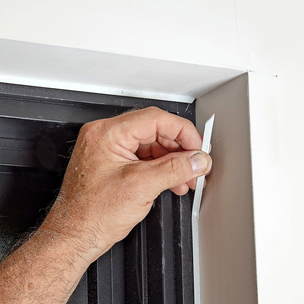 A hand holds a small, flat tool against a corner edge, smoothing a surface in a light-colored room with a dark window frame nearby.