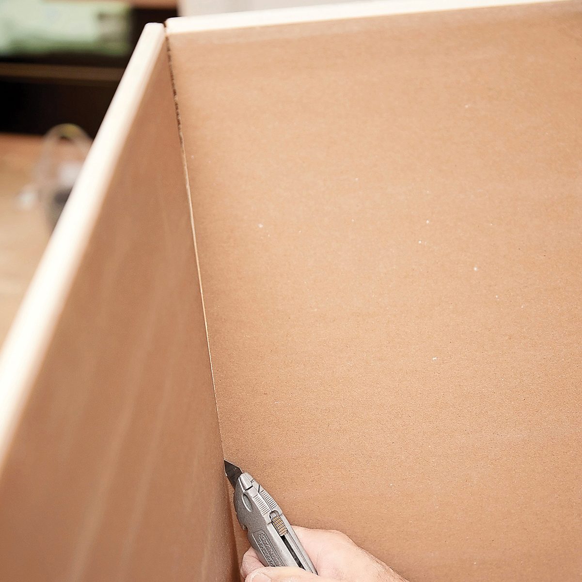A hand holds a utility knife, cutting along the seam of a cardboard box in a well-lit workshop environment.