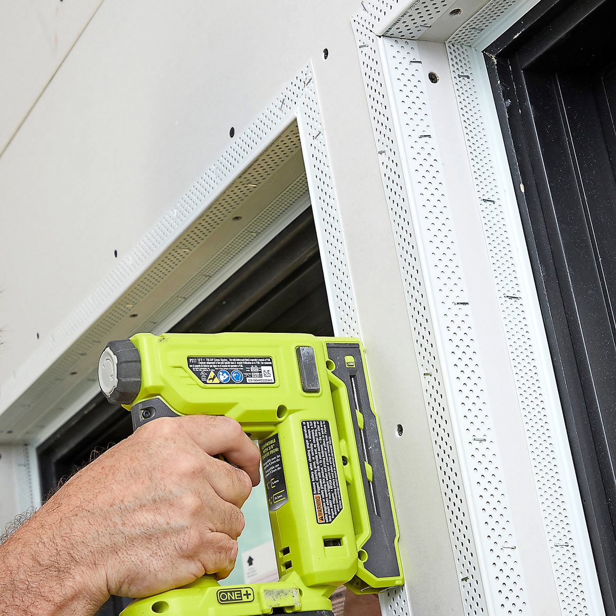 A hand operates a green power tool, securing a white frame around a window while adjacent surfaces are covered in drywall, suggesting construction or renovation.