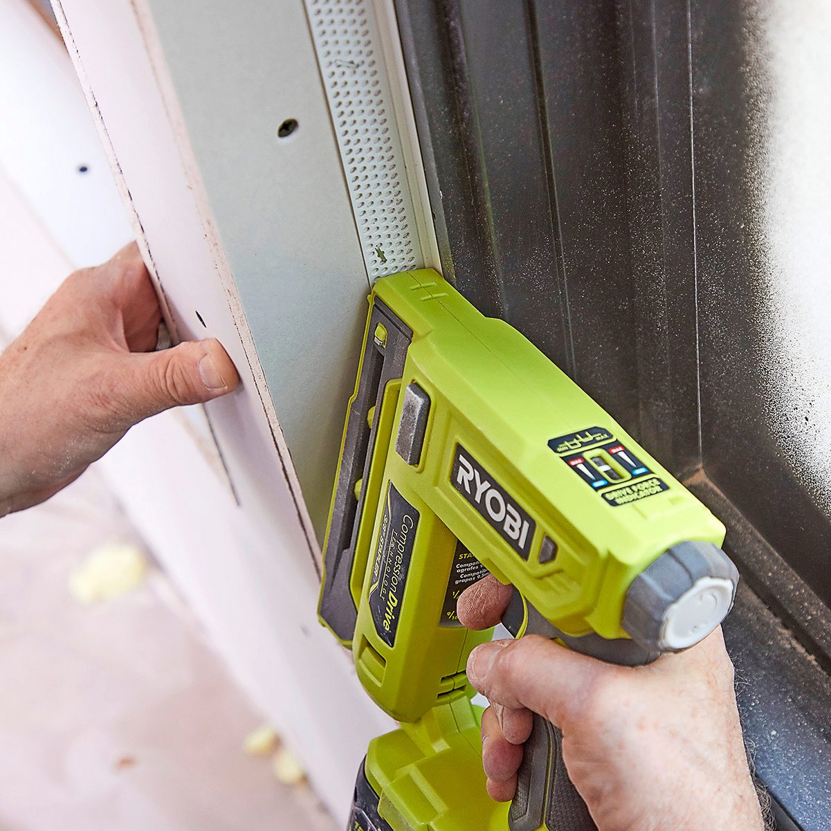 A person is using a green Ryobi power tool to attach drywall in a construction setting, with their hand pressing against the surface for support.