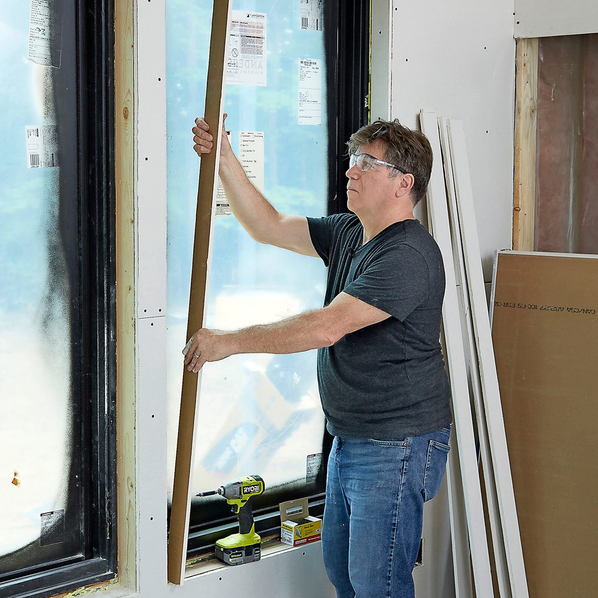 A man installs a wood panel near a window in a partially constructed room, using a power drill and surrounded by construction materials.