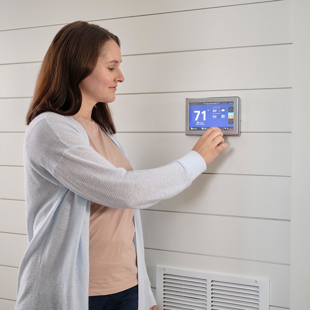 A woman adjusts a digital thermostat mounted on a light-colored wooden wall, while a vent is visible near the floor in a home environment.