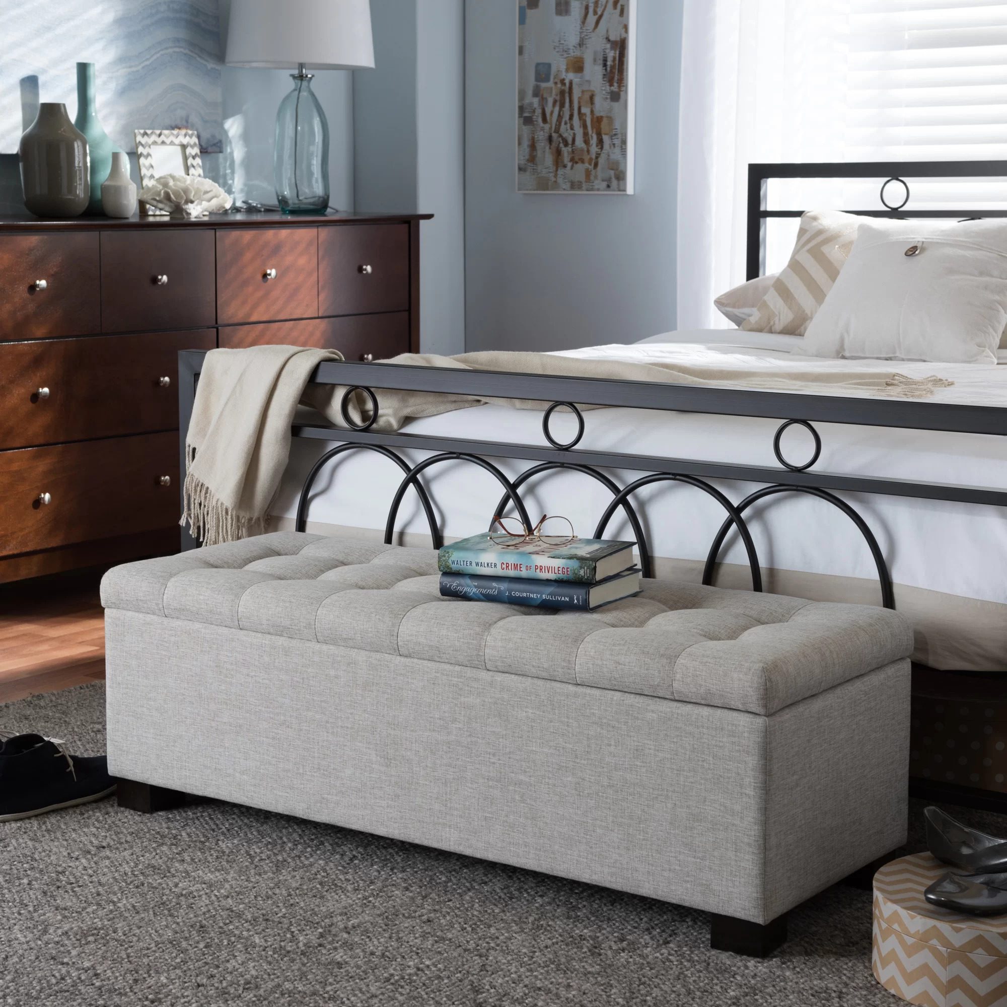 A tufted storage bench sits at the foot of a bed, holding books and glasses, amidst a softly decorated room with a dresser and lamp.