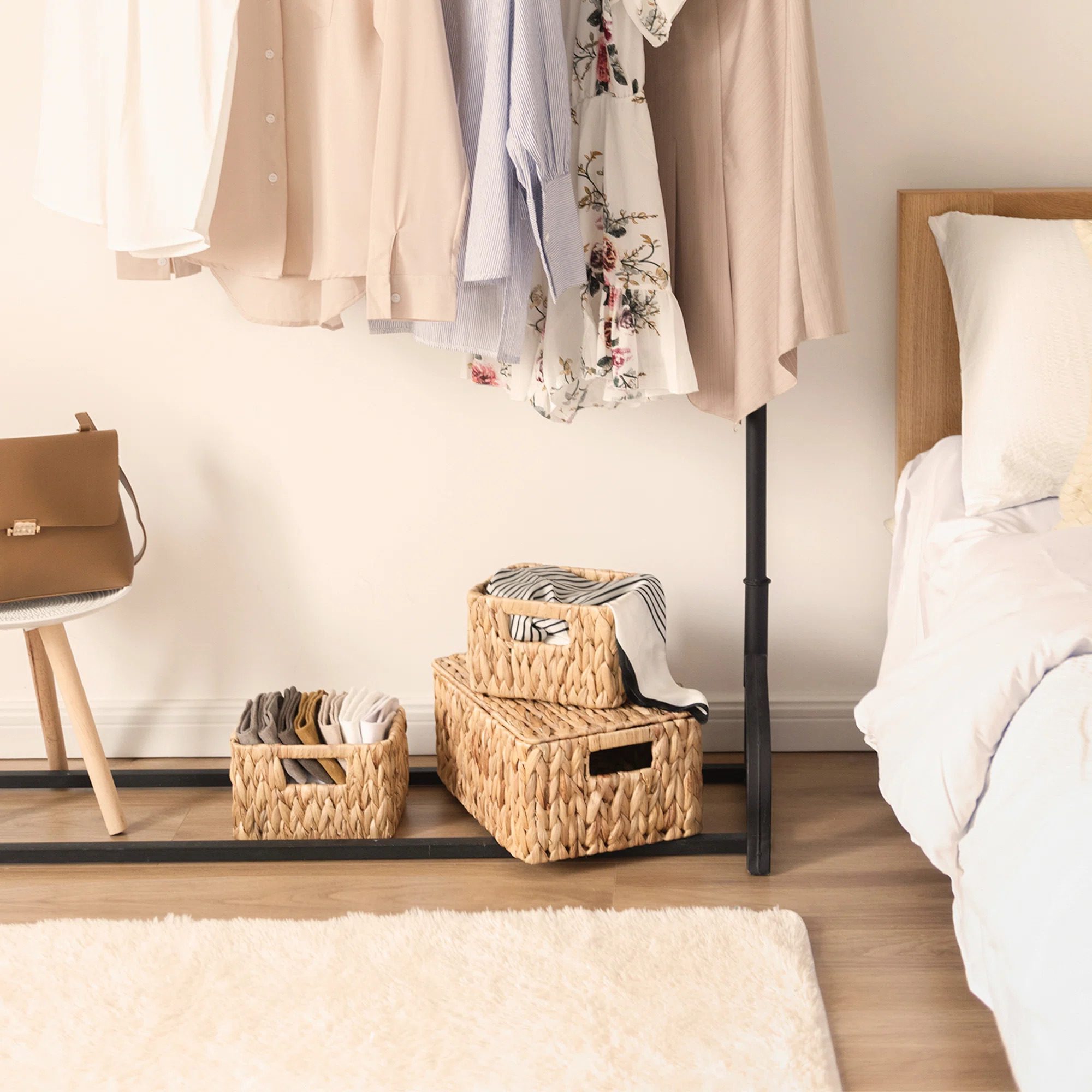 A clothing rack holds hanging shirts, while woven baskets contain folded clothes beside a chair and a bed in a softly lit bedroom.