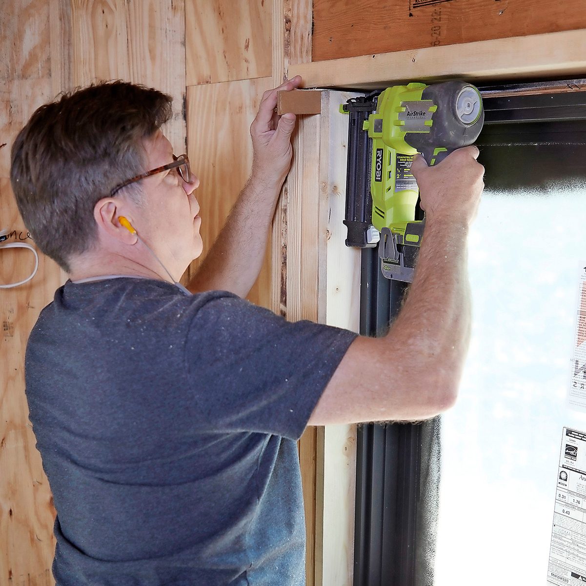 A man uses a nail gun to secure a piece of wood near a window, surrounded by wooden walls and construction materials.
