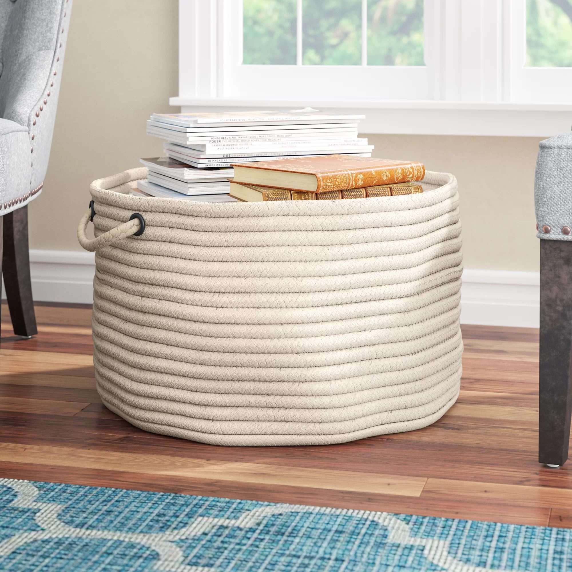 A round, fabric storage basket holds stacks of books and magazines beside a chair, on a wooden floor near a window, with a patterned rug underneath.