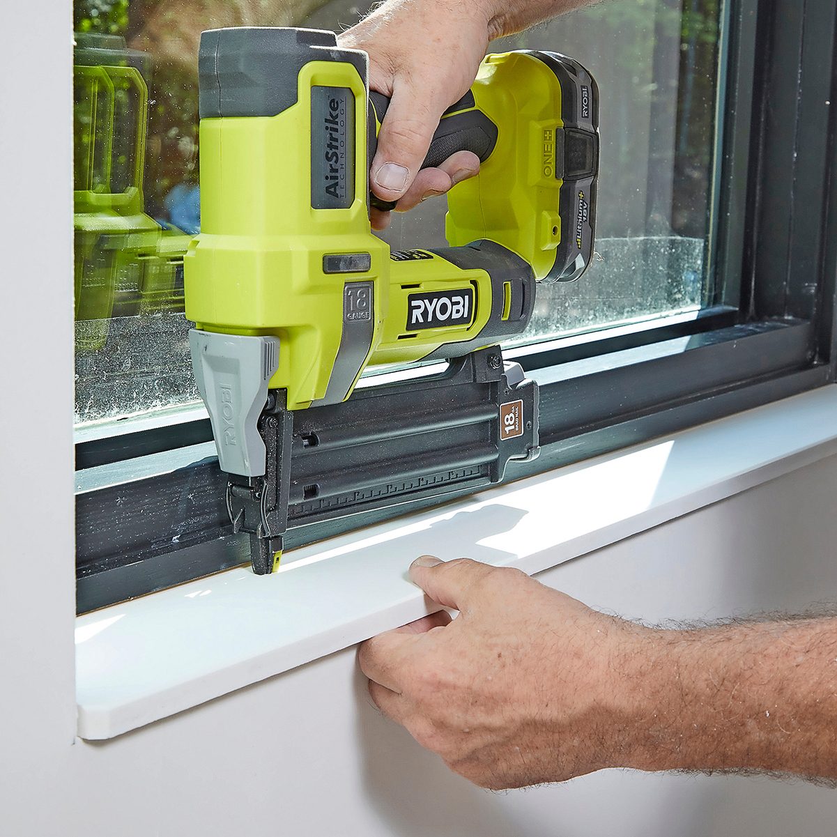 A hand operates a Ryobi nail gun, securing a white trim piece along a window ledge, with sunlight illuminating the workspace.