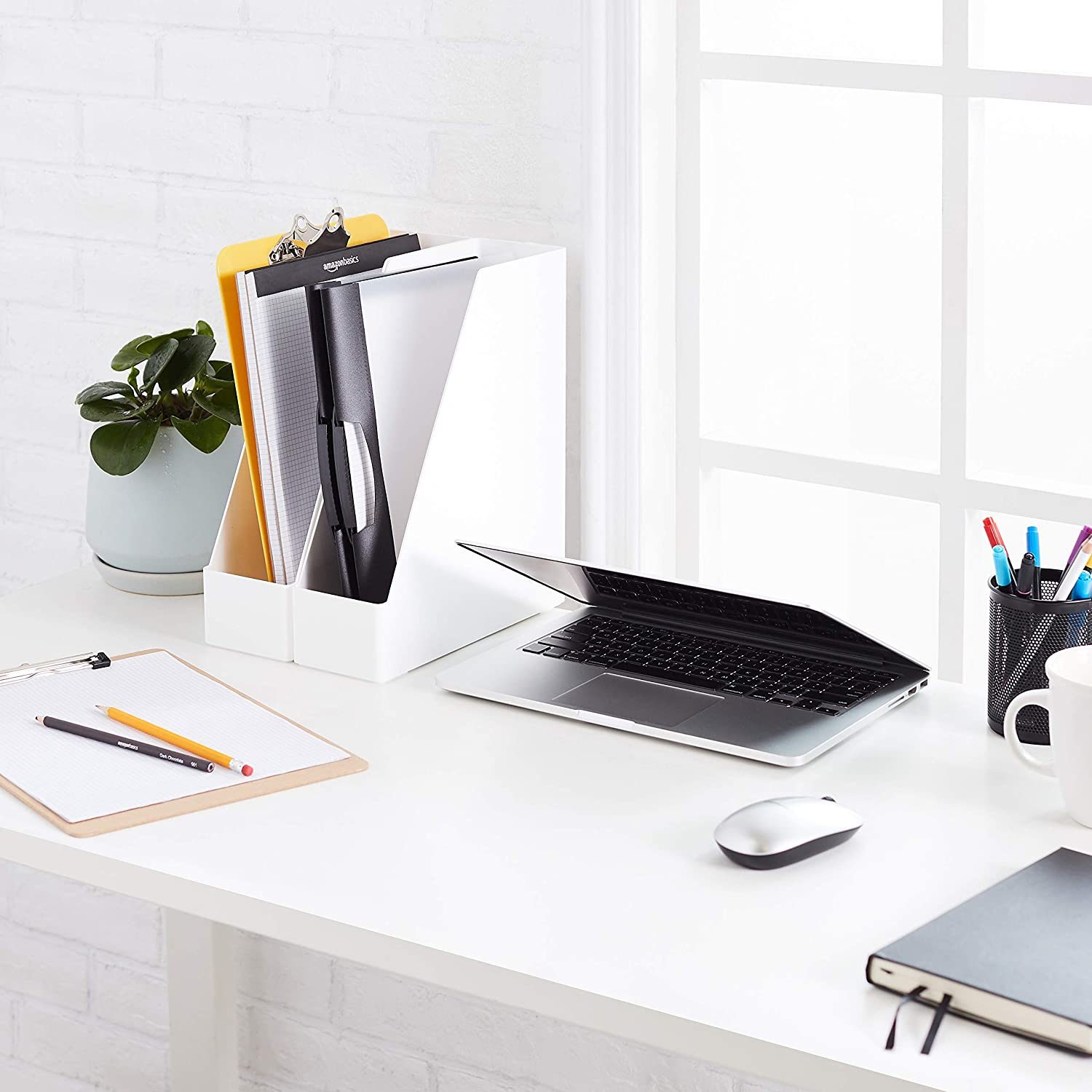 A laptop rests open on a white desk, surrounded by office supplies, a potted plant, and a window, creating a bright workspace atmosphere.