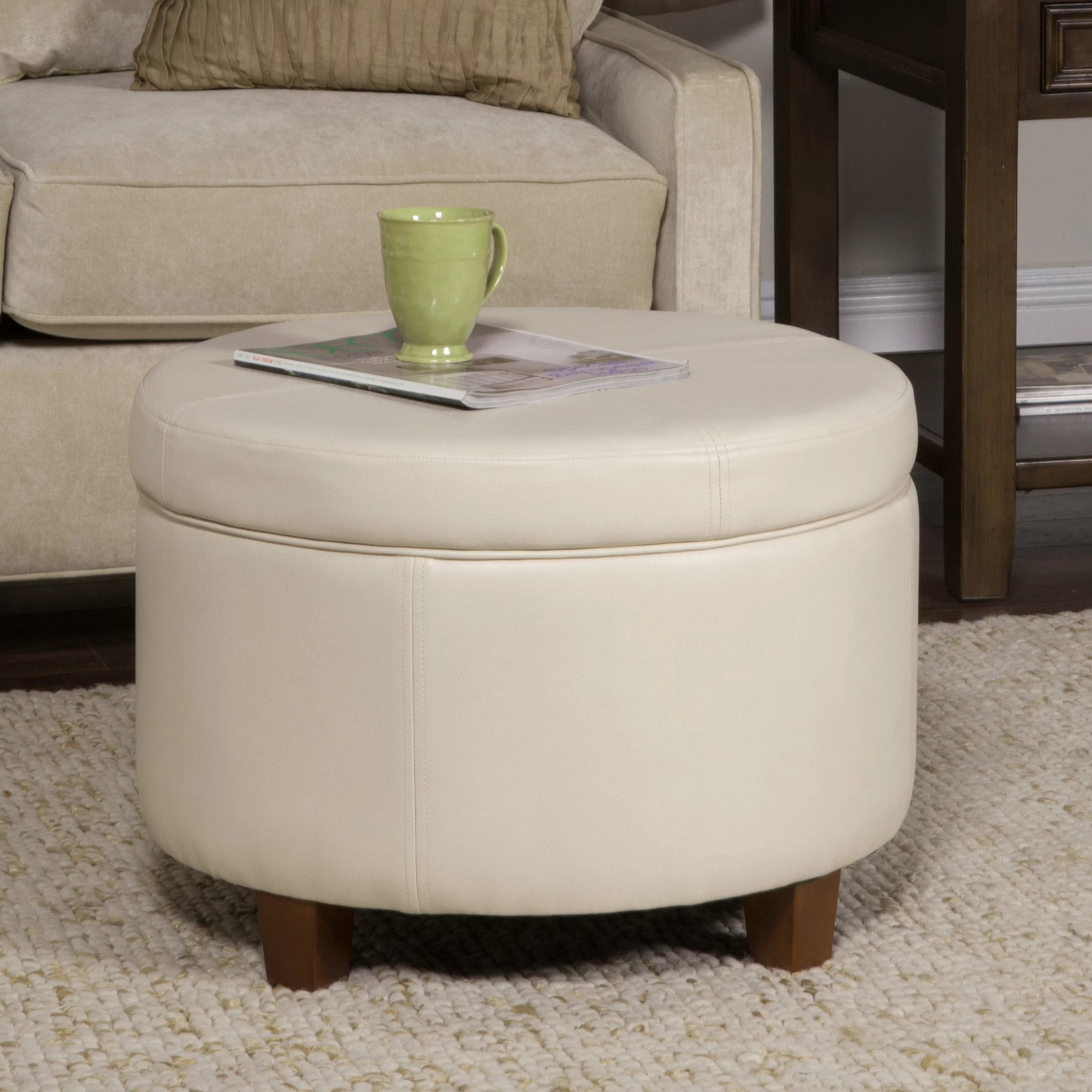 A round, cream-colored ottoman holds a green mug and a magazine, positioned in a cozy living room setting with a nearby sofa and decorative rug.