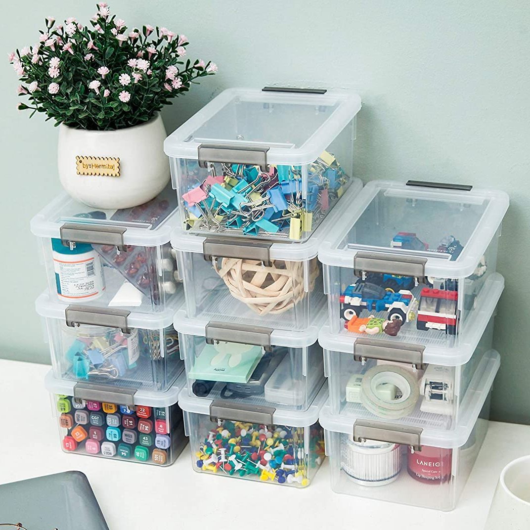 Stacked transparent storage boxes contain various office supplies and craft materials, topped with a potted plant, against a simple wall background.