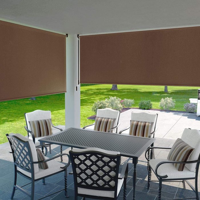 A patio table with chairs sits under brown shades. The setting is a green yard with trees and shrubs, providing a shaded outdoor dining area.
