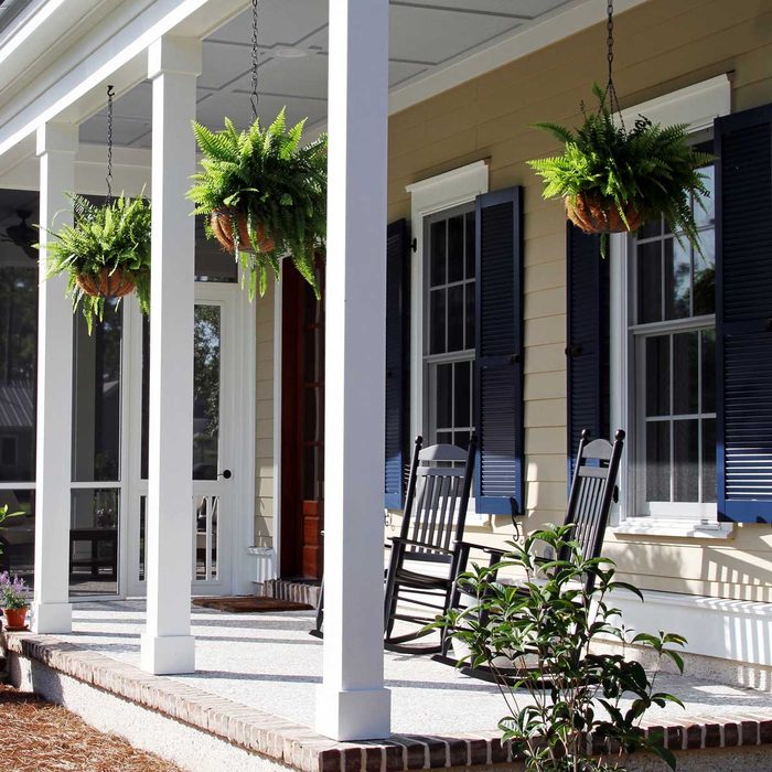 Hanging ferns sway gently above a porch with white columns, flanked by rocking chairs and framed by blue-shuttered windows on a sunny day.