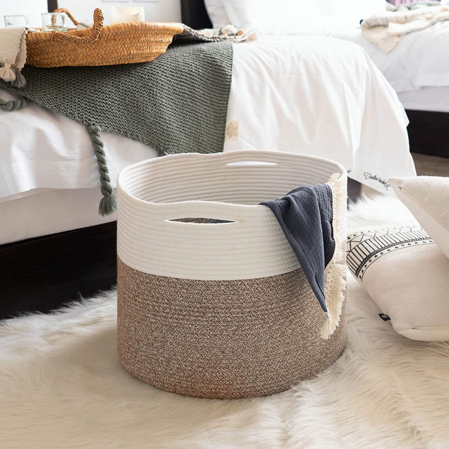 A fabric storage basket sits on a soft rug, partially filled with a dark cloth, next to a bed adorned with blankets and decorative pillows.