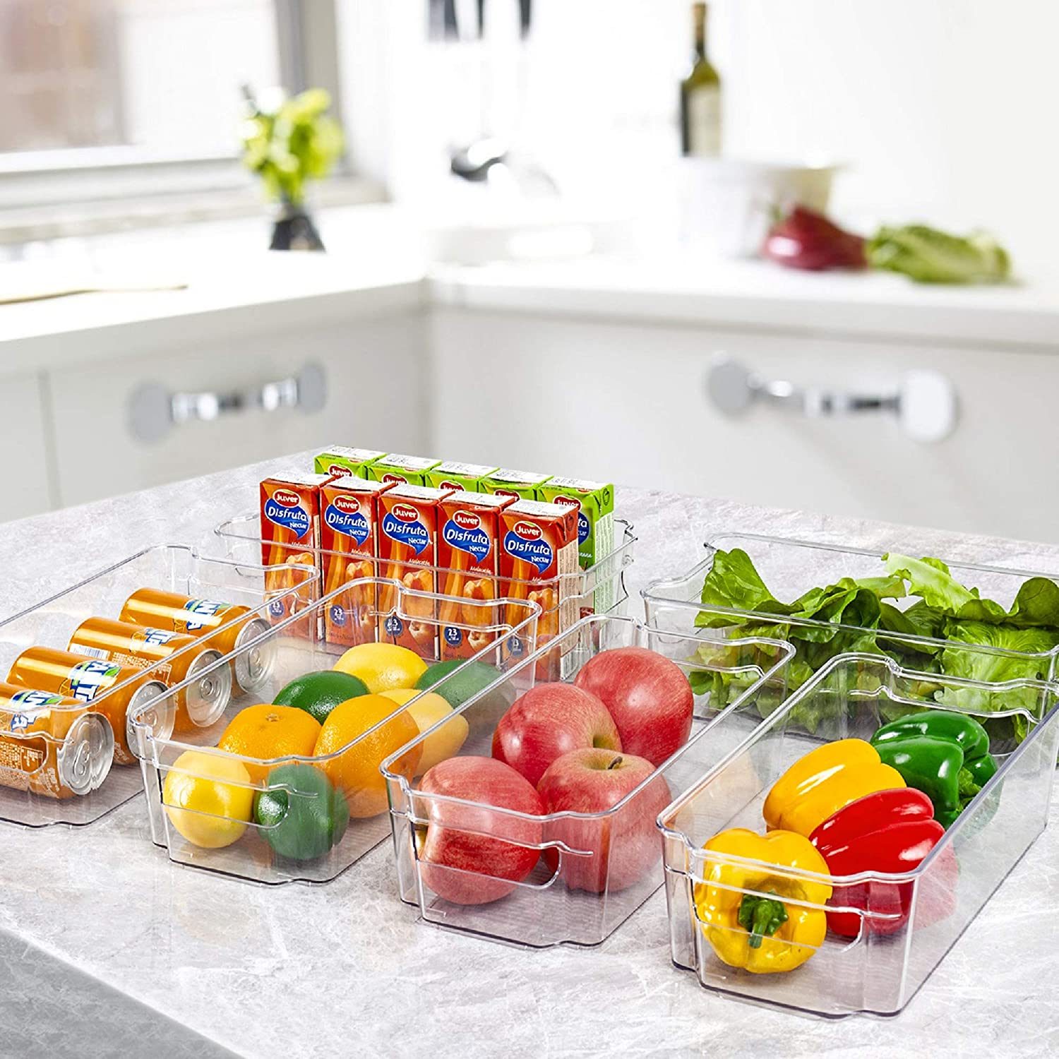 Clear containers hold assorted fruits, vegetables, and juice boxes on a kitchen countertop, surrounded by a clean, bright kitchen environment.