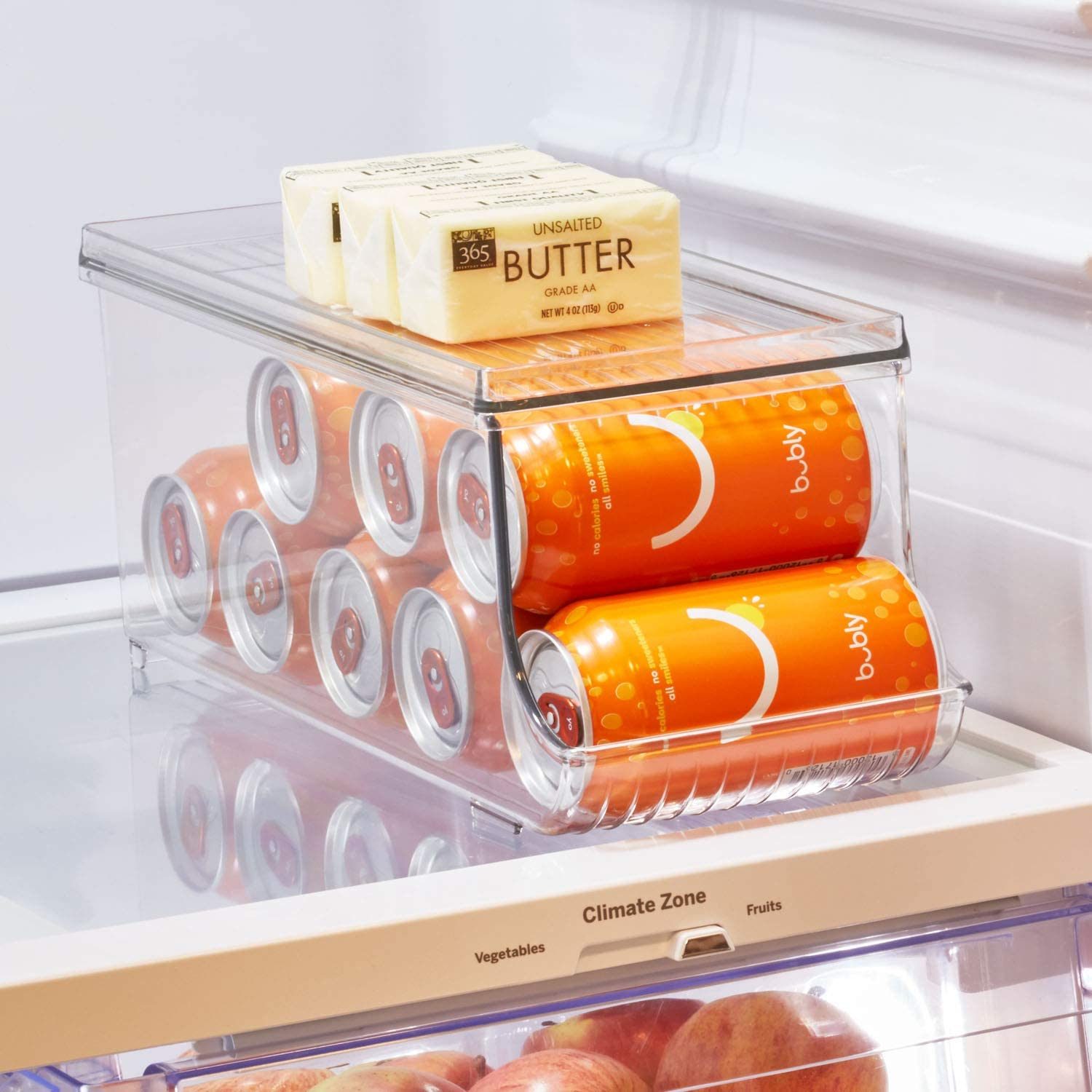 A clear refrigerator bin holds orange cans stacked upright, with butter placed on top, inside a refrigerator compartment containing apples below.