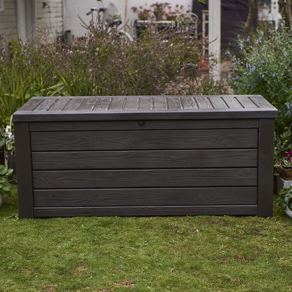A rectangular storage box rests on grass, featuring a slatted dark exterior. It's positioned among garden plants and flowers in a residential outdoor setting.