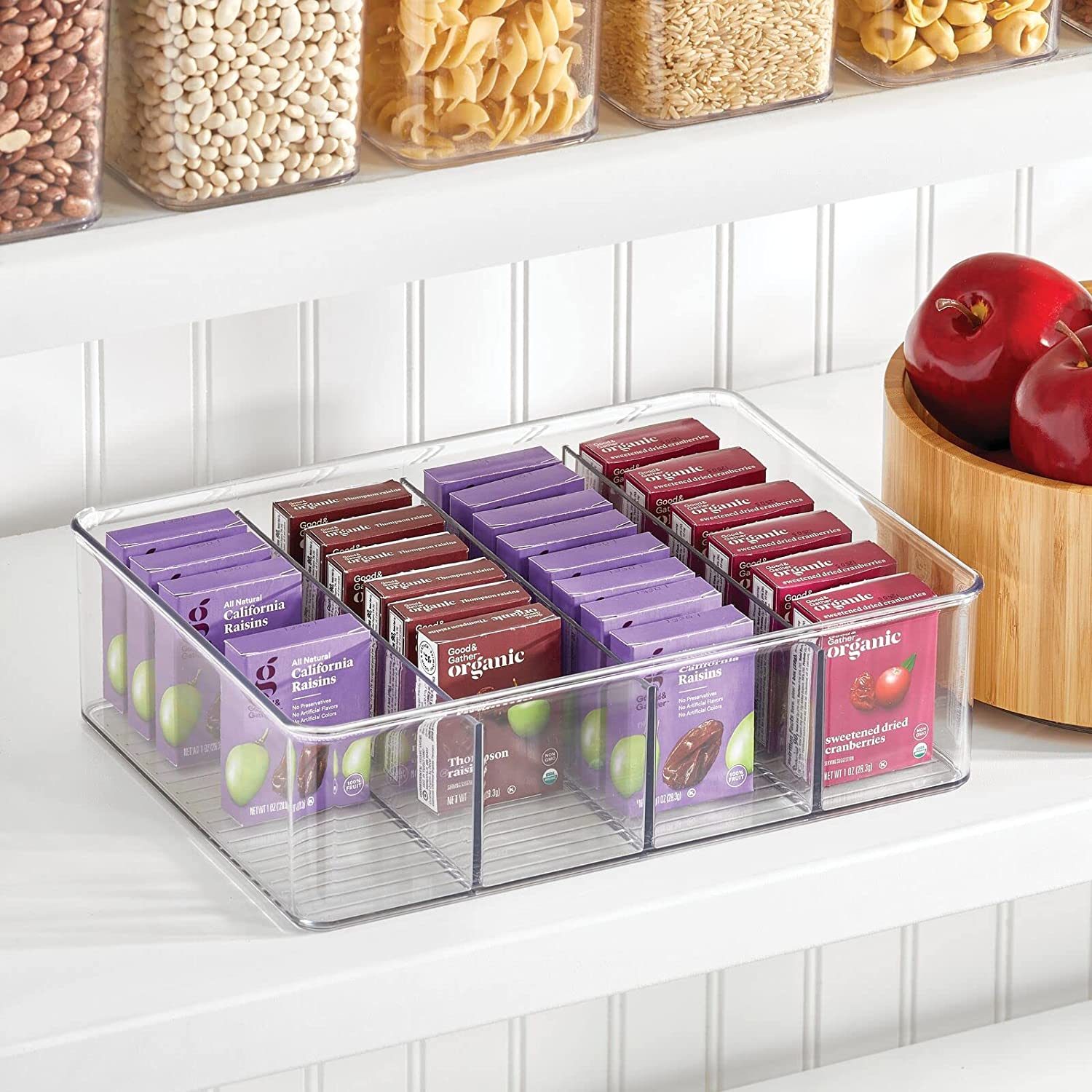 A clear container holds organized snack boxes on a shelf, with grains in jars above and a wooden bowl of red apples nearby.