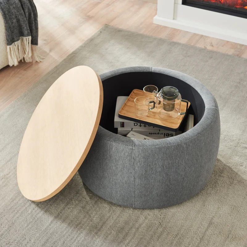 A round, upholstered ottoman with a wooden lid is open, revealing a tray, glasses, and books inside, set in a cozy living room.